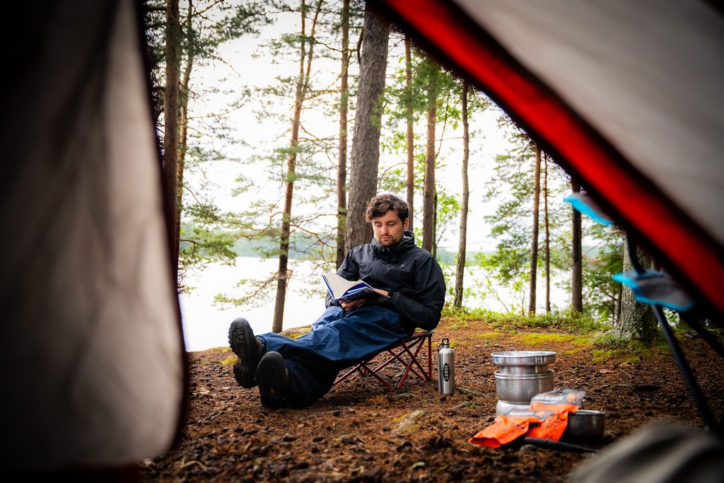 Uitzicht vanuit de tent op een man die comfortabel leest op een afgelegen wildkampeerplek aan een meer in het Zweedse Värmland, omgeven door dennenbomen.
